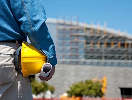 close up of hardhat and plans in a workers had with a large commercial construction site off in the distance